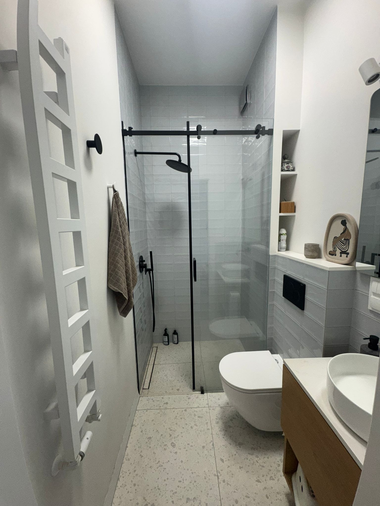 Nowoczesna łazienka po remoncie: kabina prysznicowa typu walk-in z czarnymi detalami, biała ceramika, grzejnik drabinkowy i subtelne dodatki dekoracyjne.