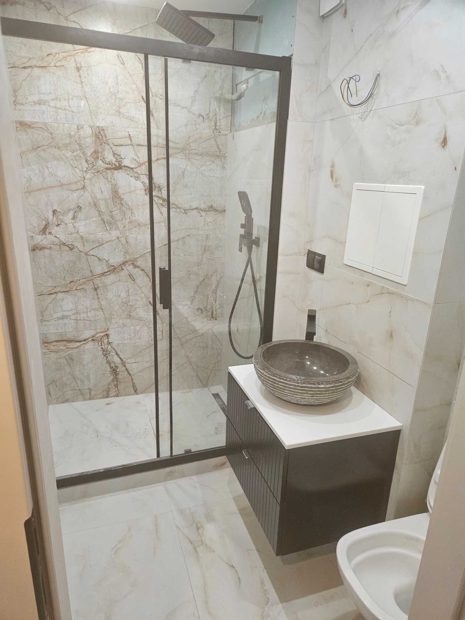 Nowoczesna łazienka z kabiną prysznicową typu walk-in, umywalką w stylu rustykalnym i marmurowymi płytkami ściennymi. Widoczne otwarte przewody elektryczne.