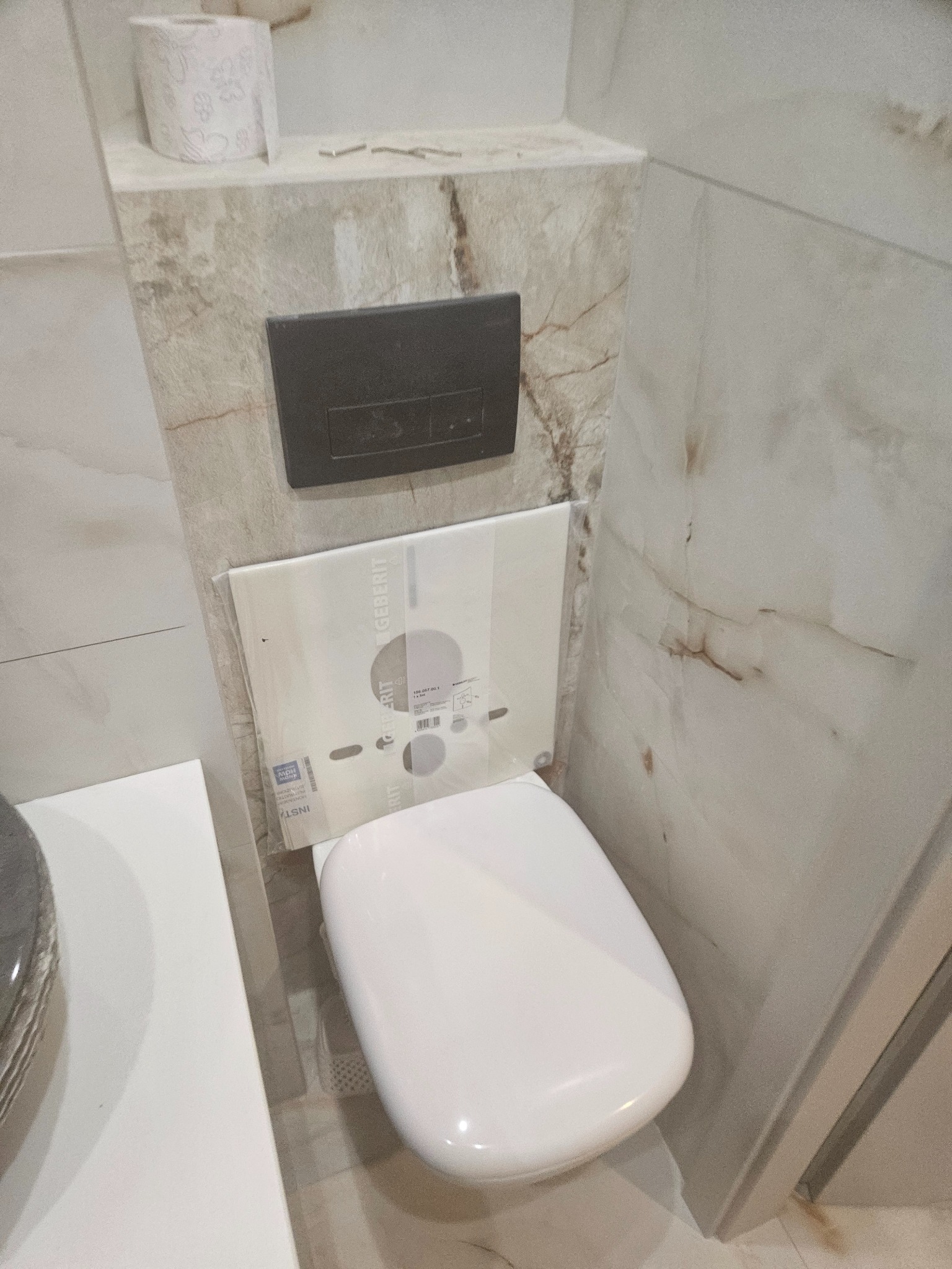 Kompakt WC z podtynkową spłuczką Geberit, obudowany płytkami imitującymi marmur. Widoczny dozownik mydła i rolka papieru toaletowego. Wykończenie łazienki w odcieniach beżu.