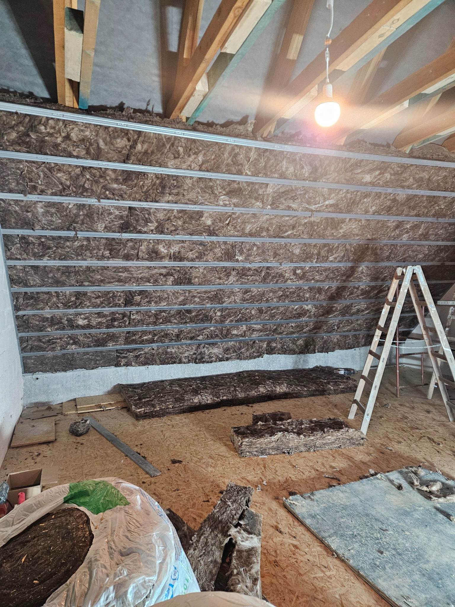 Izolacja poddasza z wełny mineralnej, widoczne metalowe profile, drabina i narzędzia. Prace w toku, surowe wykończenie.