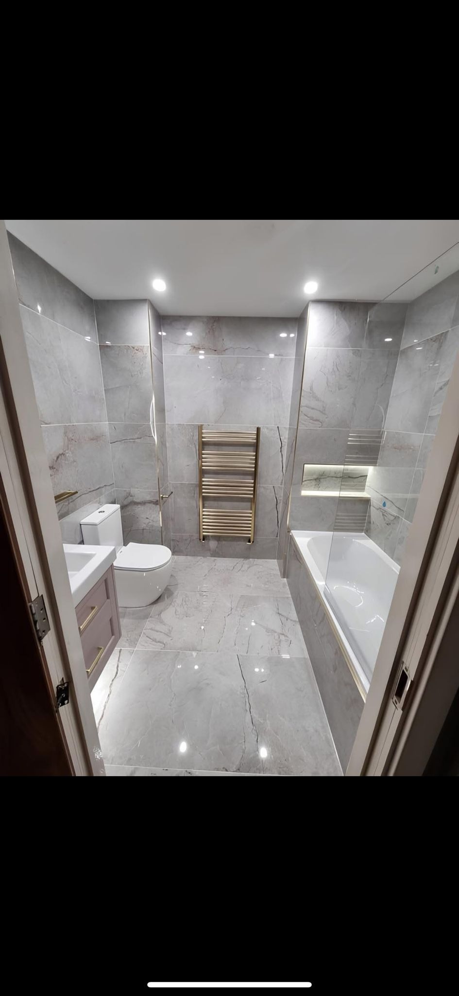 Wykończona łazienka z szarymi płytkami imitującymi marmur, złoty grzejnik drabinkowy i podświetlana wnęka nad wanną, widziana zza uchylonych drzwi.