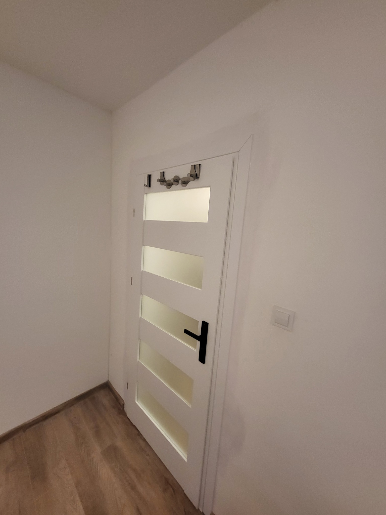 Białe drzwi wewnętrzne z poziomymi przeszkleniami i czarną klamką, zamontowane w białej ścianie. Na górze drzwi zamontowane wieszaki.