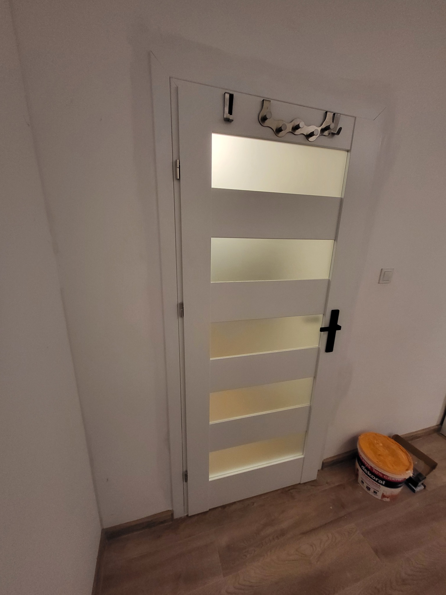 Białe drzwi z przeszkleniami i czarną klamką, na górze wieszak na ubrania. Obok ściany stoi wiadro z farbą. Podłoga drewniana.