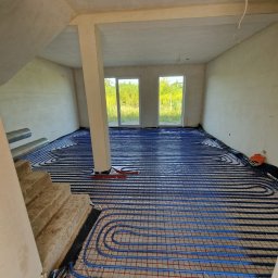 WIESŁAW SUSZEK USŁUGI WIELOBRANŻOWE - Niebieskie rury ogrzewania podłogowego ułożone na folii w surowym wnętrzu z betonowymi schodami i widokiem na zieleń przez okno.