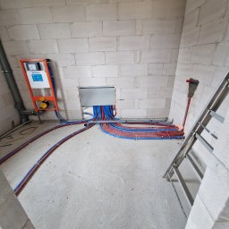 WIESŁAW SUSZEK USŁUGI WIELOBRANŻOWE - Instalacja wodna i kanalizacyjna w surowym wnętrzu: liczne rury (czerwone i niebieskie) prowadzone wzdłuż ściany z białego betonu komórkowego, widoczna drabina.