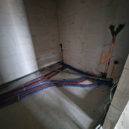 WIESŁAW SUSZEK USŁUGI WIELOBRANŻOWE - Instalacja rur grzewczych (czerwone) i wodnych (niebieskie) w surowym wnętrzu z betonu komórkowego. Widoczne pionowe wyprowadzenia rur w ścianie.