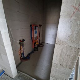 WIESŁAW SUSZEK USŁUGI WIELOBRANŻOWE - Wnęka z instalacją podtynkową WC, widoczne rury doprowadzające wodę i stelaż. Ściany z surowych bloczków, podłoga wylewka. Prace hydrauliczne w trakcie realizacji.