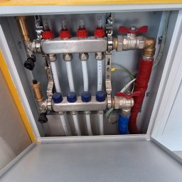 WIESŁAW SUSZEK USŁUGI WIELOBRANŻOWE - Rozdzielacz ogrzewania podłogowego w metalowej szafce z otwartymi drzwiczkami, widoczne białe rury i zawory z czerwonymi i niebieskimi elementami regulacyjnymi.