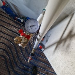 WIESŁAW SUSZEK USŁUGI WIELOBRANŻOWE - Instalacja ogrzewania podłogowego z widocznym manometrem i zaworami, ułożona na siatce. Ujęcie z góry, widoczny fragment ściany i but.