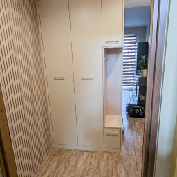 Antoni Wajdzik Mevalo - Beżowa szafa wnękowa z siedziskiem i złotymi uchwytami w przedpokoju z panelami ściennymi i drewnopodobną podłogą. Widok z perspektywy wejścia do pomieszczenia.