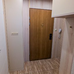 Antoni Wajdzik Mevalo - Beżowa szafa wnękowa z frontami bez uchwytów, drewniane panele ścienne i drzwi wejściowe z ozdobnym wizjerem w kształcie podkowy tworzą spójną aranżację przedpokoju.