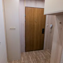 Beżowa szafa wnękowa z frontami bez uchwytów, drewniane panele ścienne i drzwi wejściowe z ozdobnym wizjerem w kształcie podkowy tworzą spójną aranżację przedpokoju.