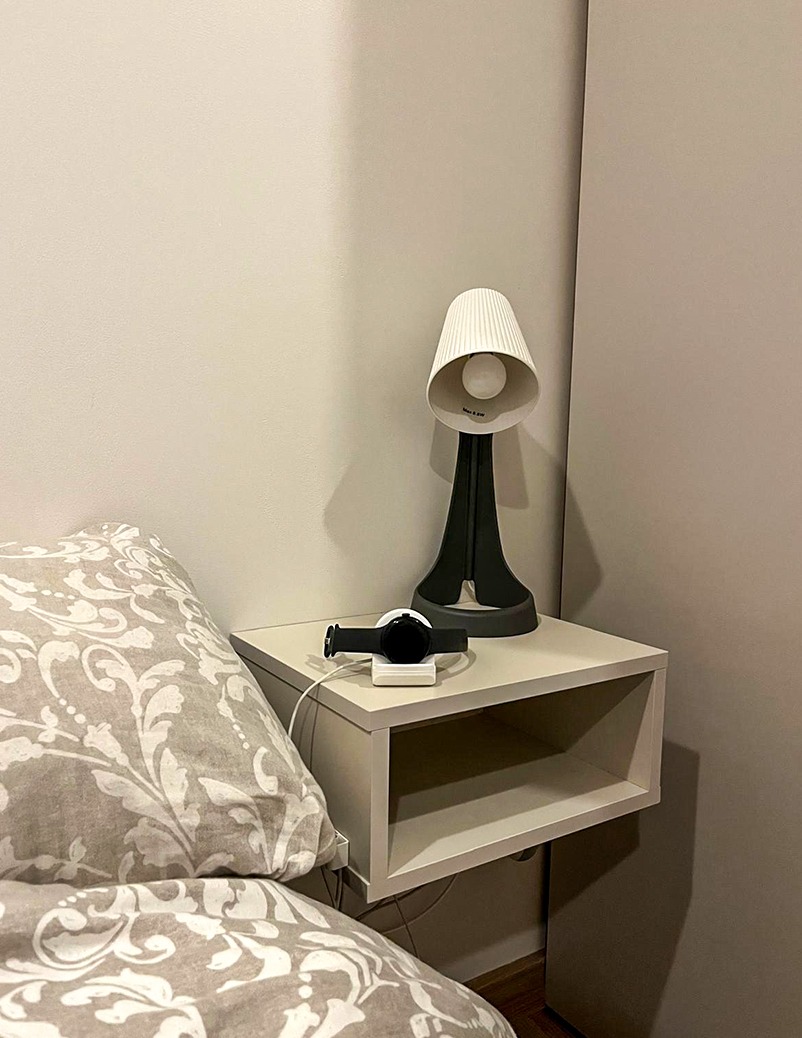 Biała szafka nocna z lampką i zegarkiem, przymocowana do ściany obok łóżka z wzorzystą pościelą. Minimalistyczny design, ciepłe światło.