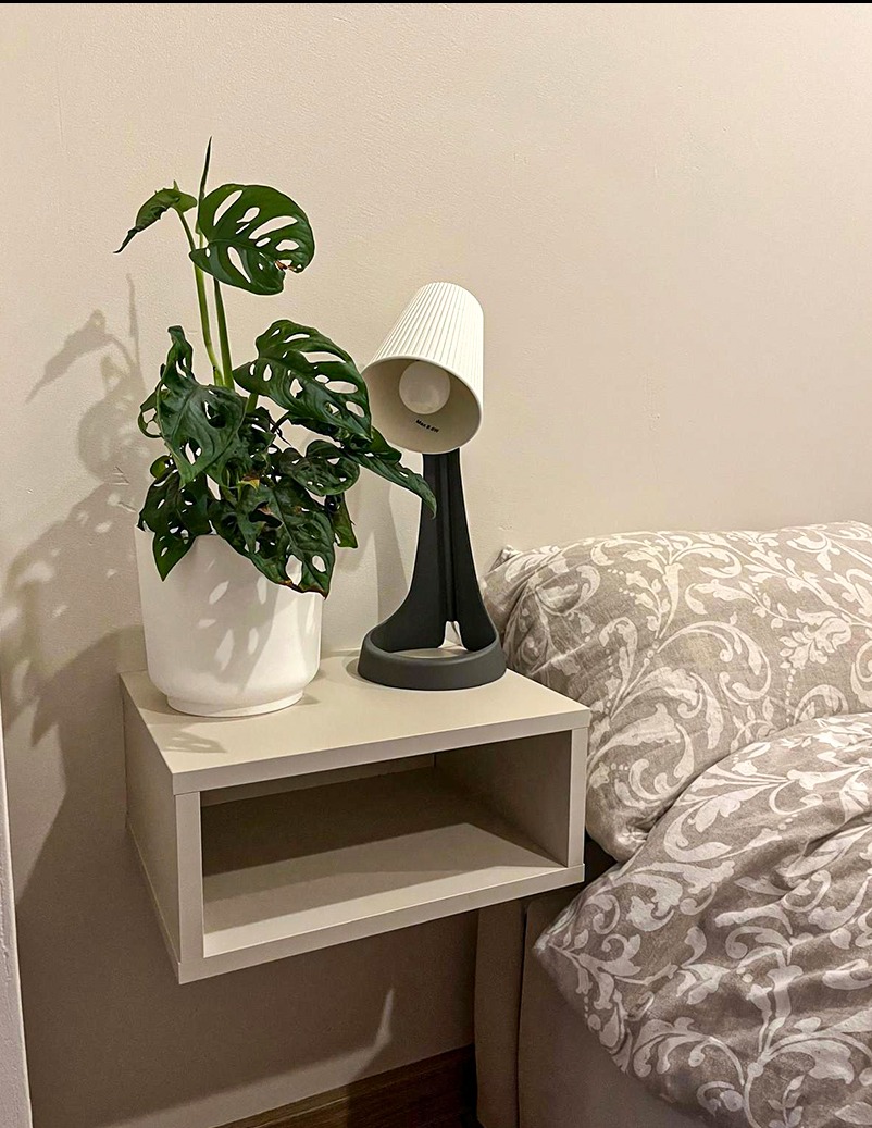 Beżowa szafka nocna wisząca z rośliną w doniczce i lampką, obok łóżko z wzorzystą pościelą. Minimalistyczny design, ciepłe oświetlenie.