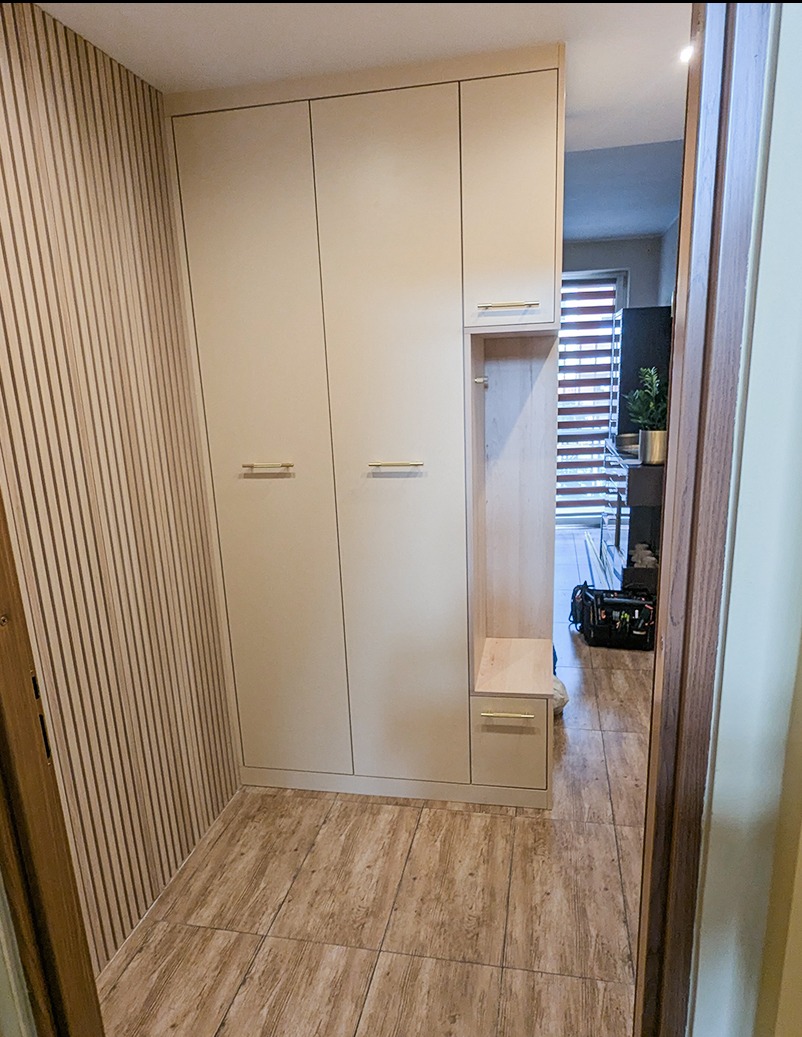 Beżowa szafa wnękowa z siedziskiem i złotymi uchwytami w przedpokoju z panelami ściennymi i drewnopodobną podłogą. Widok z perspektywy wejścia do pomieszczenia.