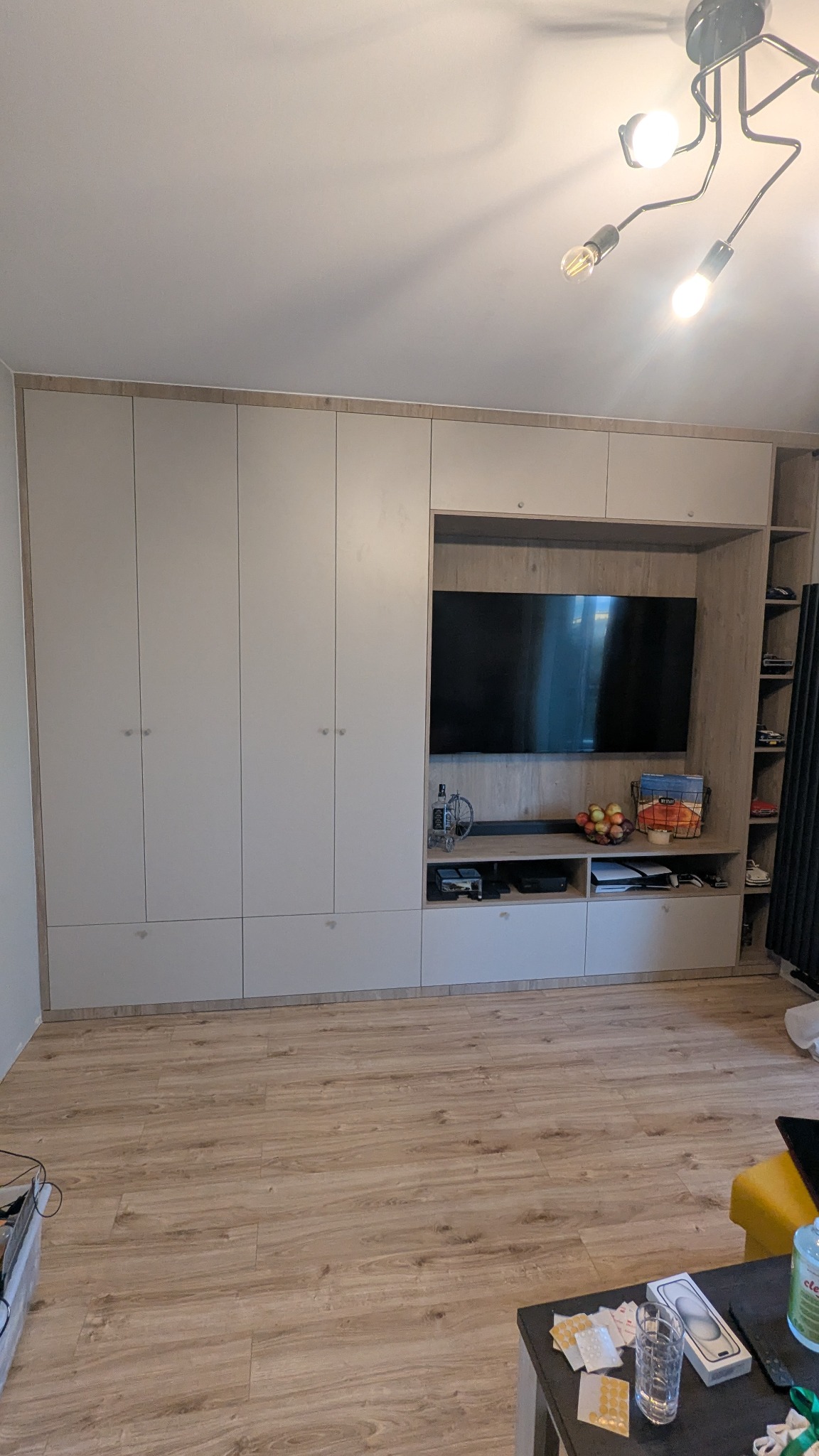 Jasna zabudowa meblowa z szafami i miejscem na TV, zintegrowana z półkami i szafkami. Widoczny fragment podłogi i nowoczesna lampa sufitowa.