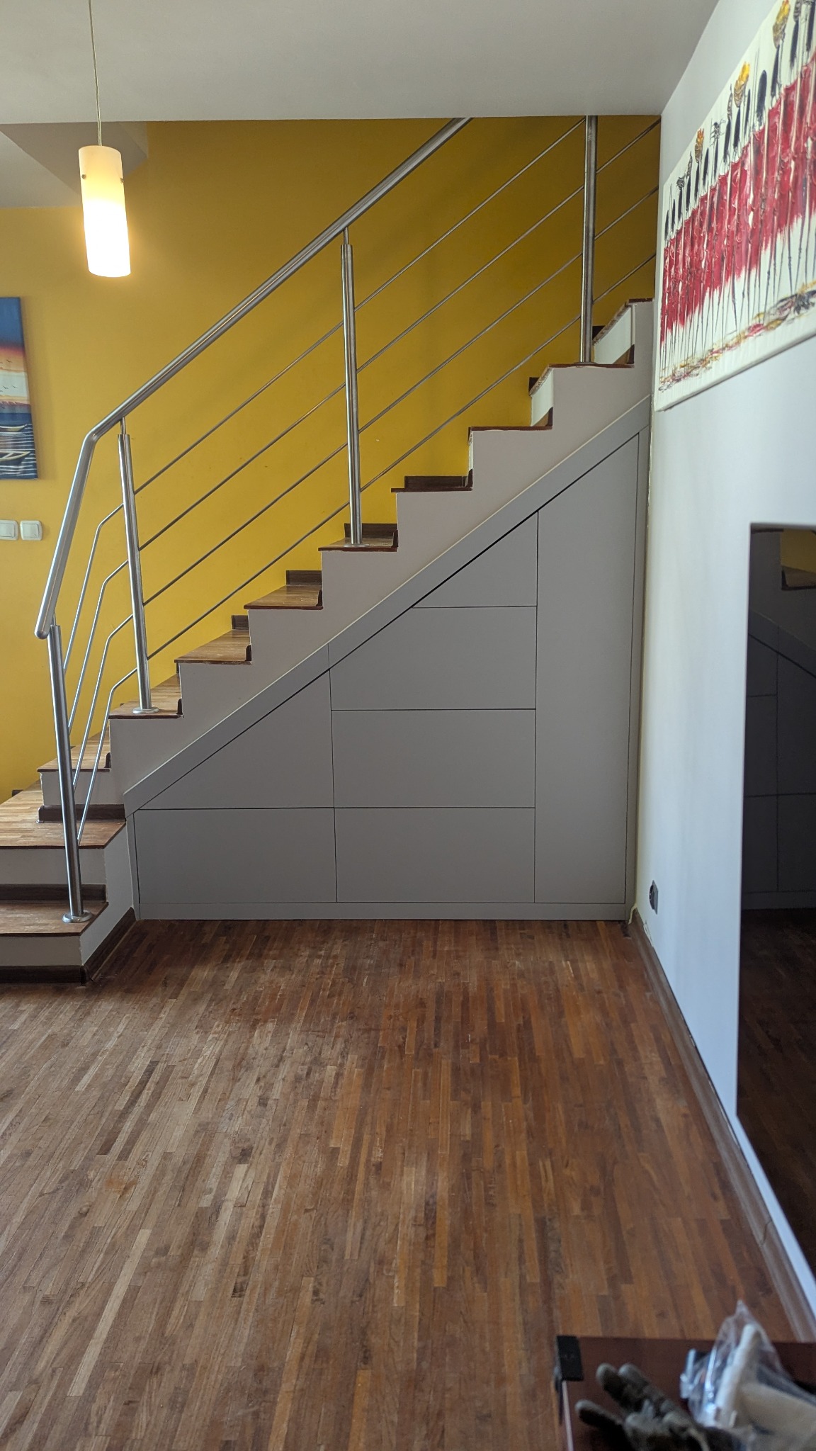 Szafa wnękowa pod schodami z drewnianymi stopniami i metalową balustradą na tle żółtej ściany. Widoczna drewniana podłoga i obraz na ścianie.