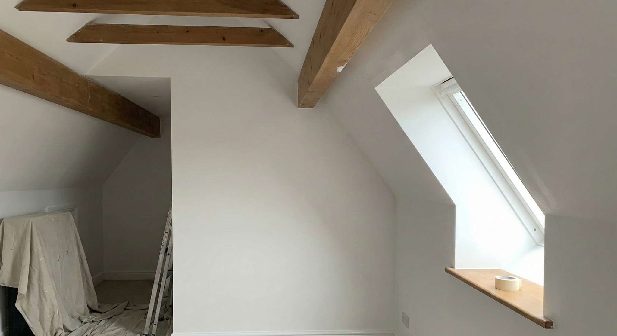Poddasze po malowaniu na biało, z drewnianymi belkami stropowymi i oknem dachowym. Na parapecie leży rolka taśmy malarskiej. Widoczna drabina i zabezpieczone meble.