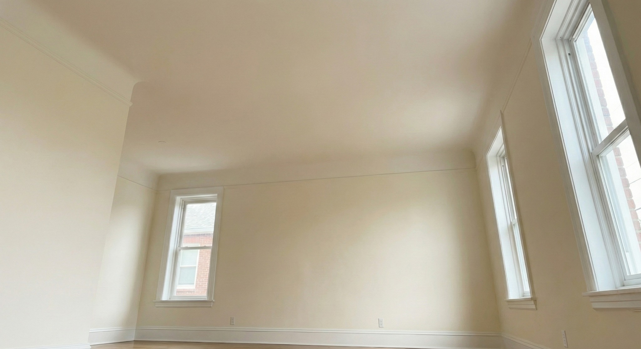 Jasne, świeżo pomalowane wnętrze pokoju z trzema oknami i drewnianą podłogą. Ściany w odcieniach beżu, białe listwy przypodłogowe i okienne. Widok z dołu, podkreślający przestrzeń.