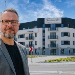 GN Nieruchomości Grzegorz Niedźwiedź - Mężczyzna w szarej marynarce na tle nowoczesnego apartamentowca z logo 'Twarowski Developer' i rondem. Prezentacja dewelopera na tle inwestycji.