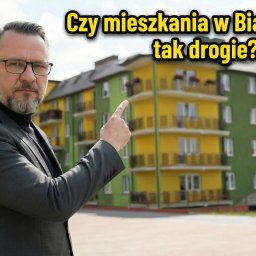 GN Nieruchomości Grzegorz Niedźwiedź - Mężczyzna w garniturze wskazuje na kolorowy budynek mieszkalny z balkonami. Tekst: Czy mieszkania w Białej są już tak drogie?