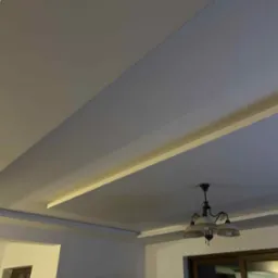 Dwupoziomowy sufit z oświetleniem LED i nowoczesną lampą wiszącą. Ściany pomalowane na dwa odcienie szarości. W tle widoczny fragment okna i rośliny.