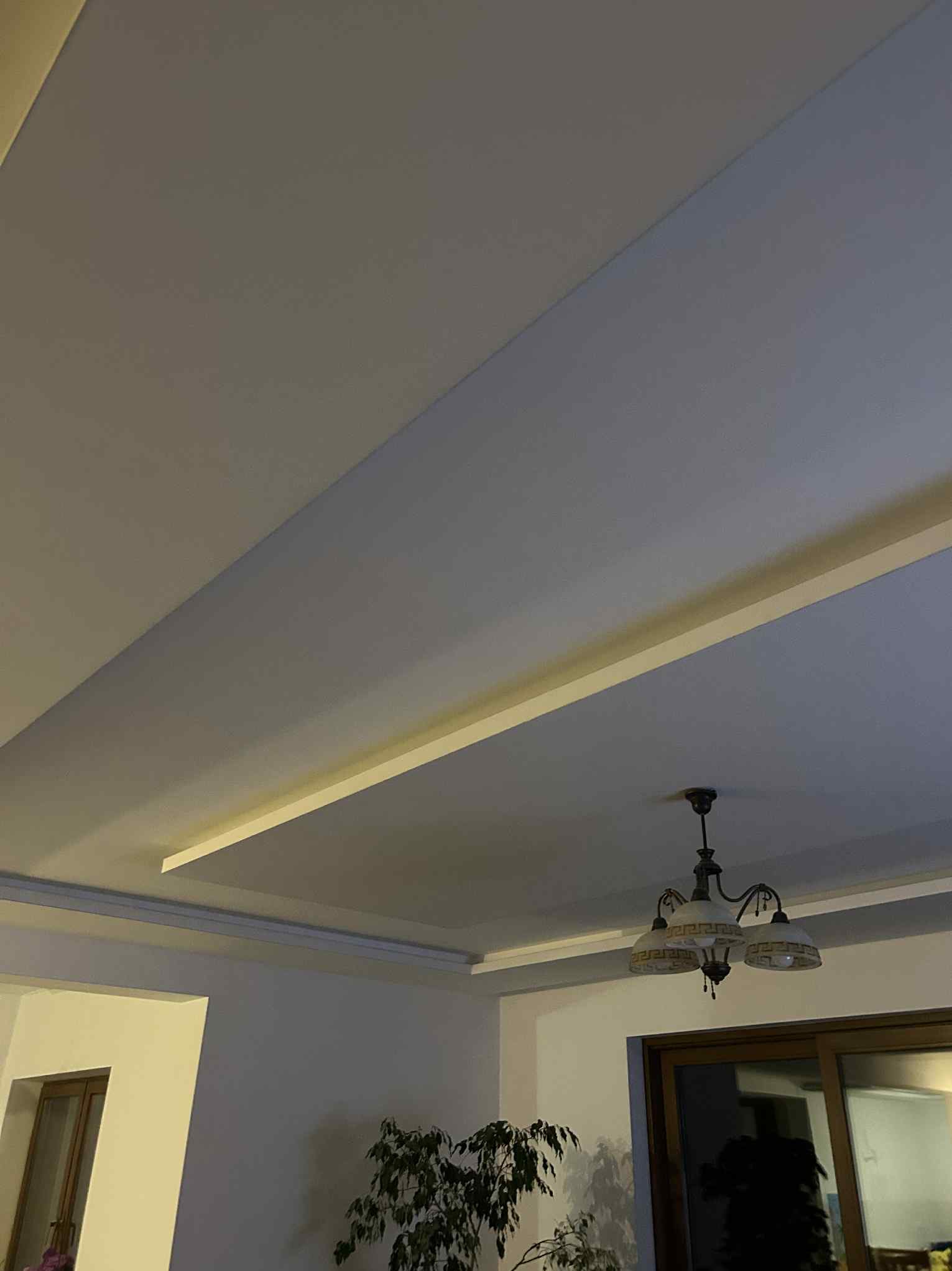 Dwupoziomowy sufit z oświetleniem LED i nowoczesną lampą wiszącą. Ściany pomalowane na dwa odcienie szarości. W tle widoczny fragment okna i rośliny.