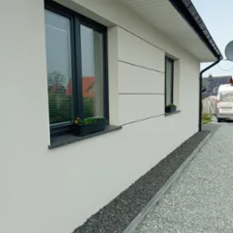 Ściana domu z oknami w ciemnych ramach i dekoracyjnymi paskami elewacyjnymi. Na parapetach kwiaty w doniczkach. Pod oknami kamienny pas oddzielający elewację od żwirowego podłoża.