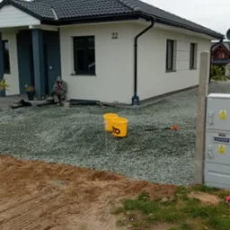 Biały dom z numerem 22, obsypany szarym żwirem. Dwa żółte wiadra i grabie leżą na żwirze. Osoba siedzi w pobliżu wejścia. W tle pochmurne niebo.