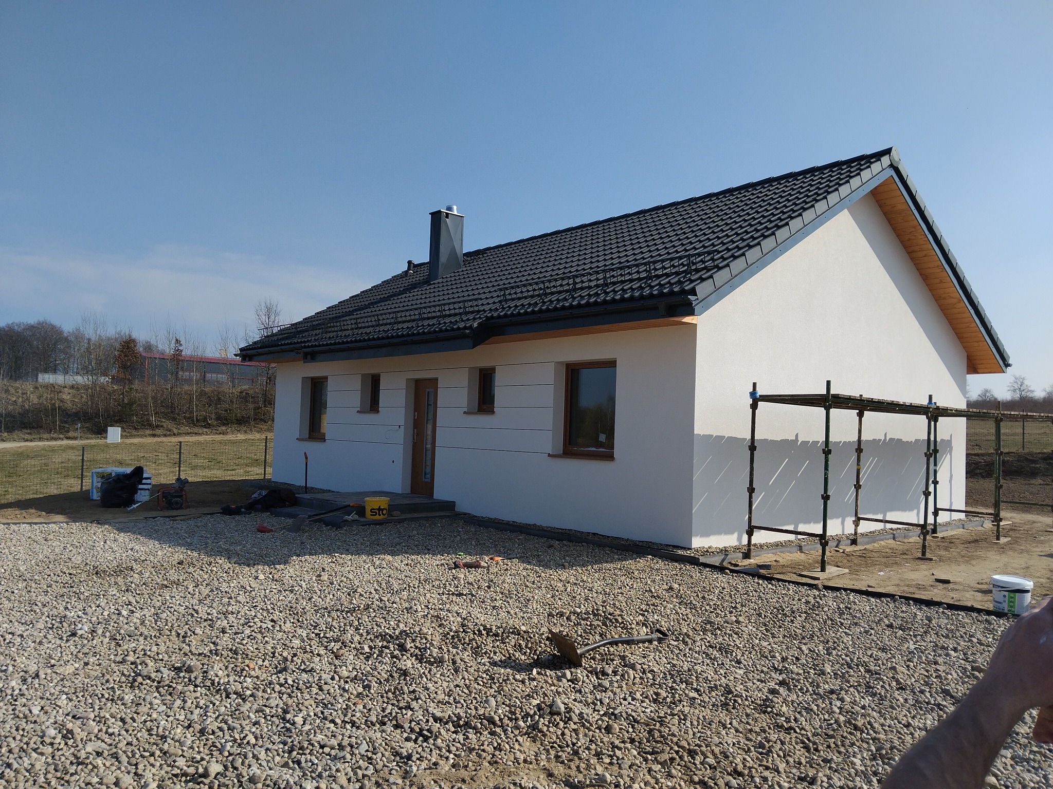 Biały dom z ciemną dachówką, widoczny z boku, otoczony żwirowym terenem. Na elewacji widoczne rusztowania. Elementy krajobrazu w tle.
