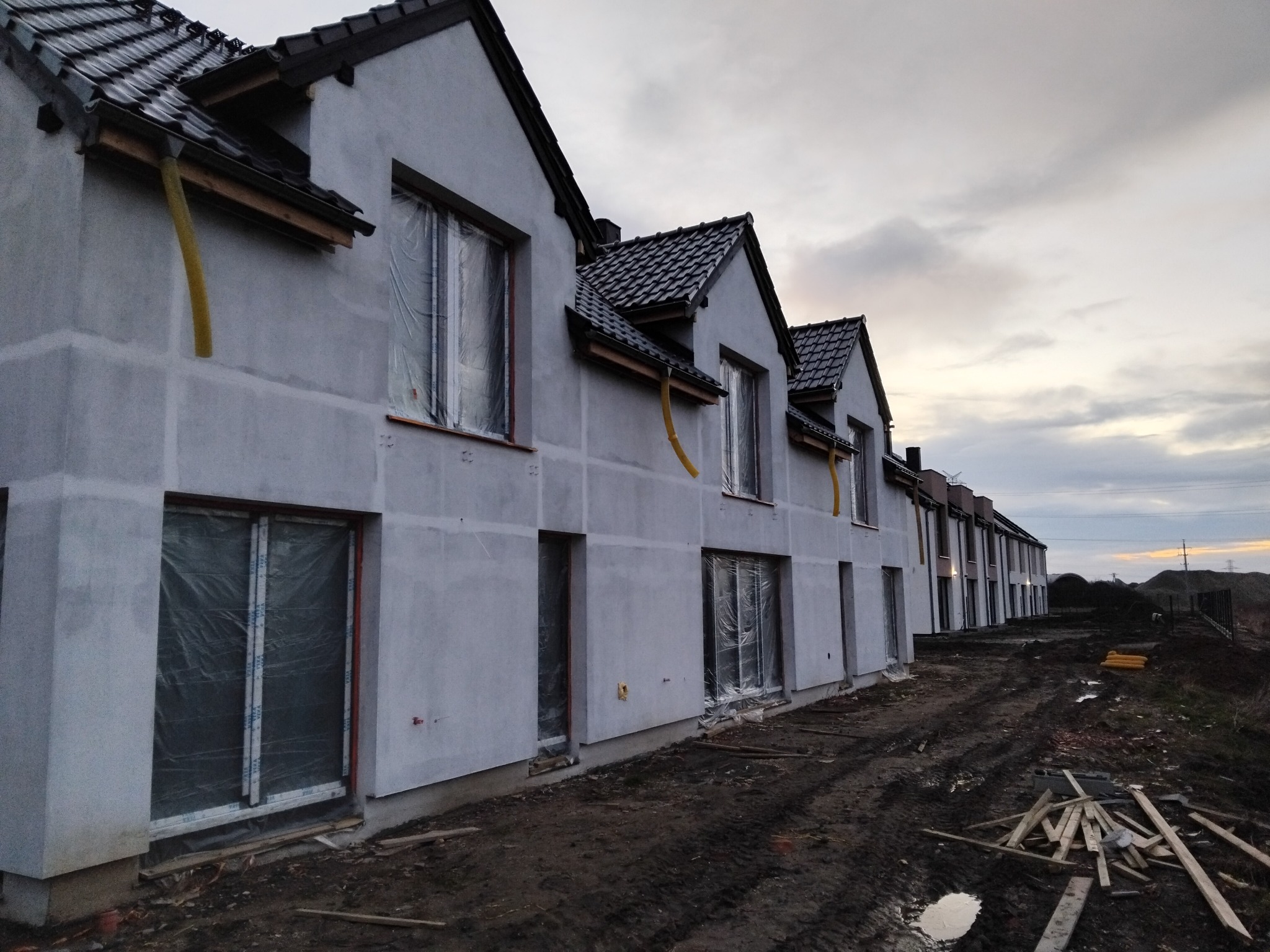 Rząd szeregowych domów w stanie surowym zamkniętym, elewacja szara, okna zabezpieczone folią, widoczne rury spustowe. Teren wokół domów błotnisty.