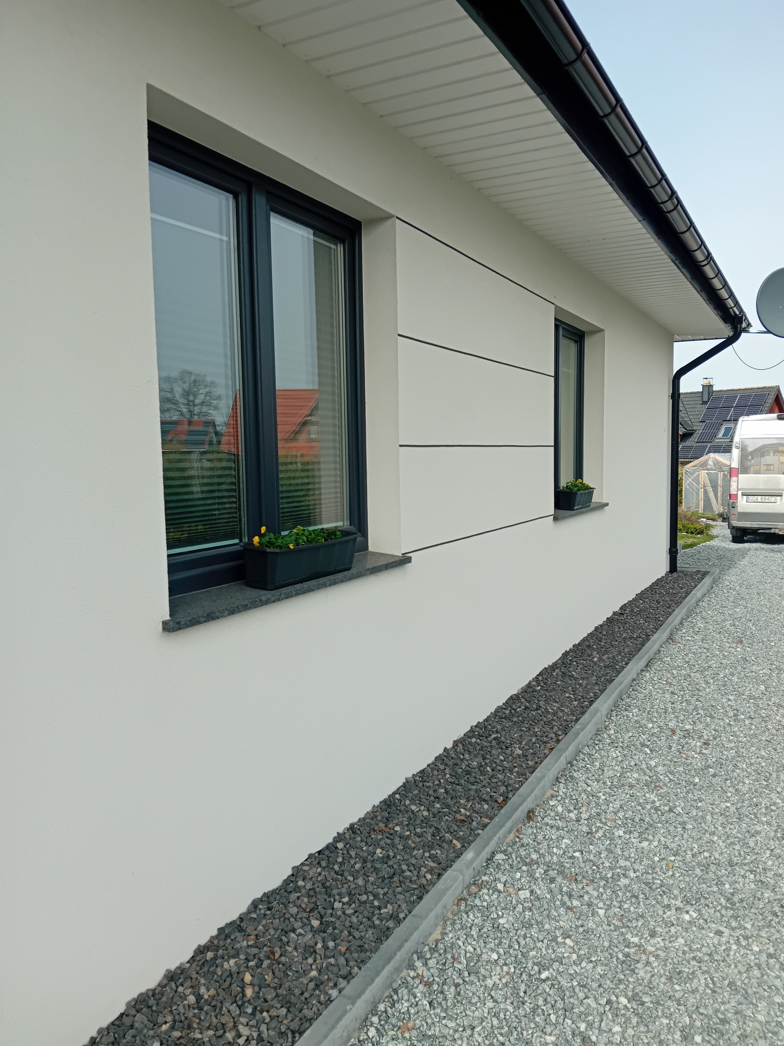 Ściana domu z oknami w ciemnych ramach i dekoracyjnymi paskami elewacyjnymi. Na parapetach kwiaty w doniczkach. Pod oknami kamienny pas oddzielający elewację od żwirowego podłoża.