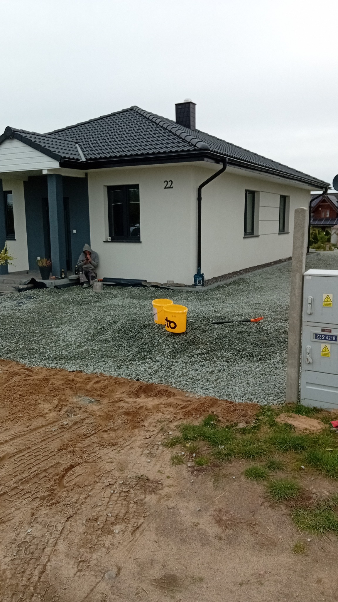 Biały dom z numerem 22, obsypany szarym żwirem. Dwa żółte wiadra i grabie leżą na żwirze. Osoba siedzi w pobliżu wejścia. W tle pochmurne niebo.