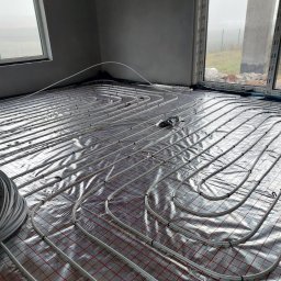 Maciej Duda - Białe rury ogrzewania podłogowego ułożone na folii z czerwoną kratką w surowym wnętrzu z oknami. Widoczny zwój rur i elementy mocujące.