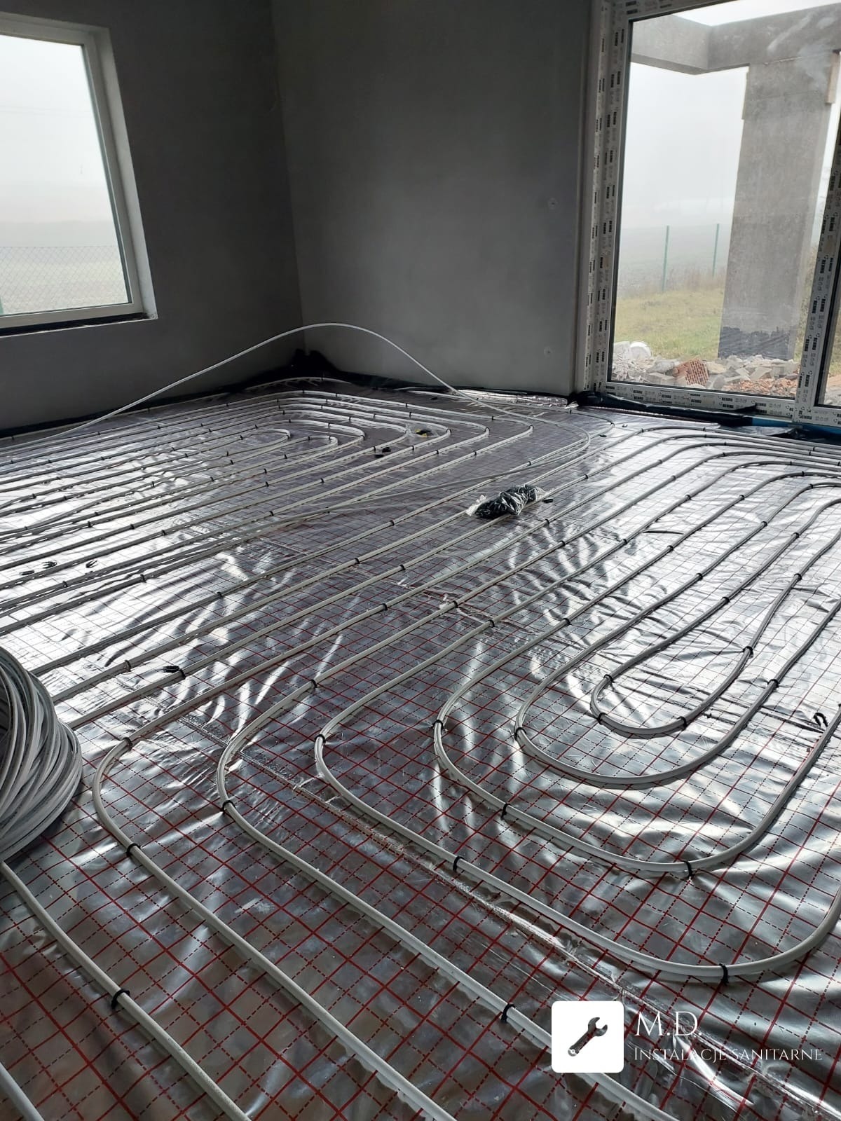 Białe rury ogrzewania podłogowego ułożone na folii z czerwoną kratką w surowym wnętrzu z oknami. Widoczny zwój rur i elementy mocujące.