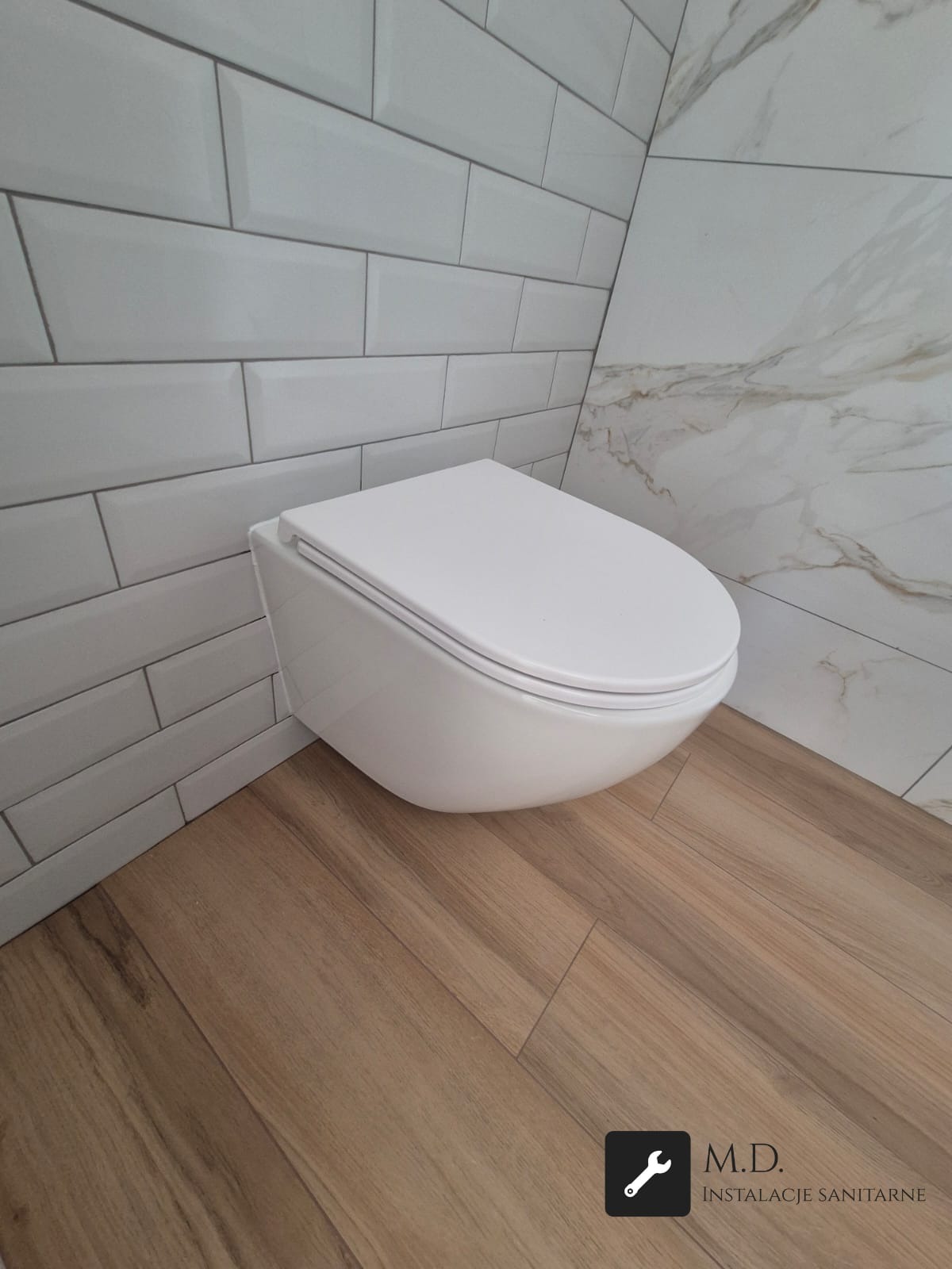 Nowoczesna, podwieszana toaleta w wykończonej łazience z białymi, cegiełkowymi płytkami i marmurową ścianą akcentującą. Parkiet imitujący drewno na podłodze.