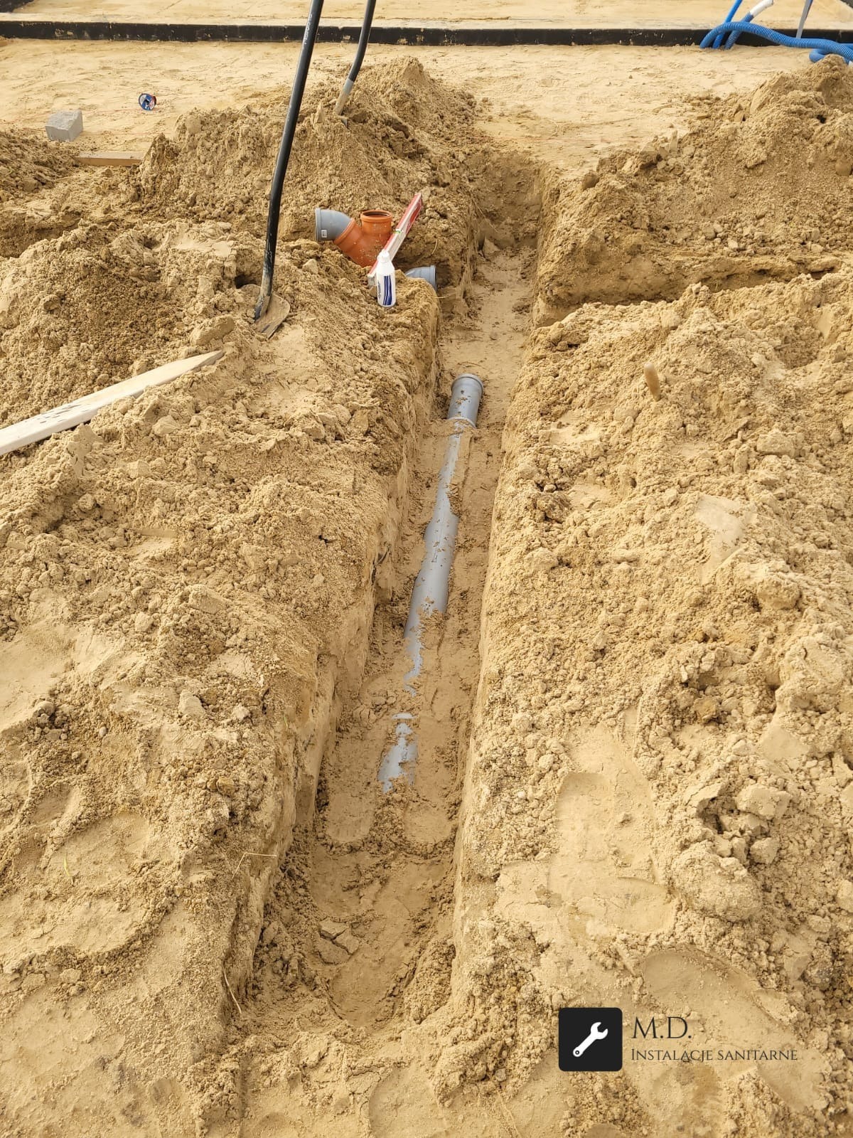 Szara rura kanalizacyjna ułożona w wykopie w piaszczystej ziemi. Widoczne narzędzia i elementy instalacji. Ujęcie z góry, ukazujące etap prac instalacyjnych.