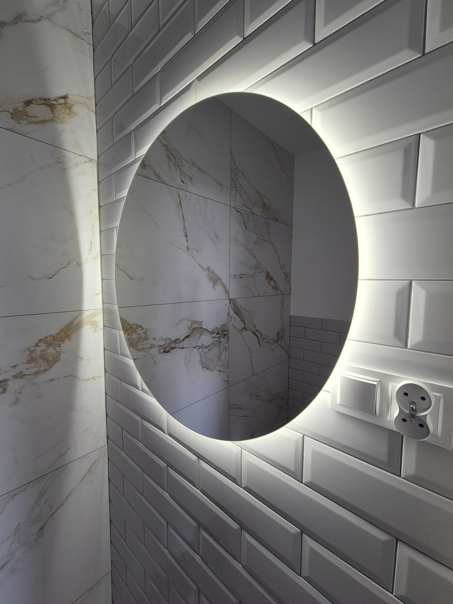 Okrągłe lustro z podświetleniem LED na ścianie wyłożonej białymi, podłużnymi płytkami. Widoczny fragment ściany z płytkami imitującymi marmur i gniazdko elektryczne.