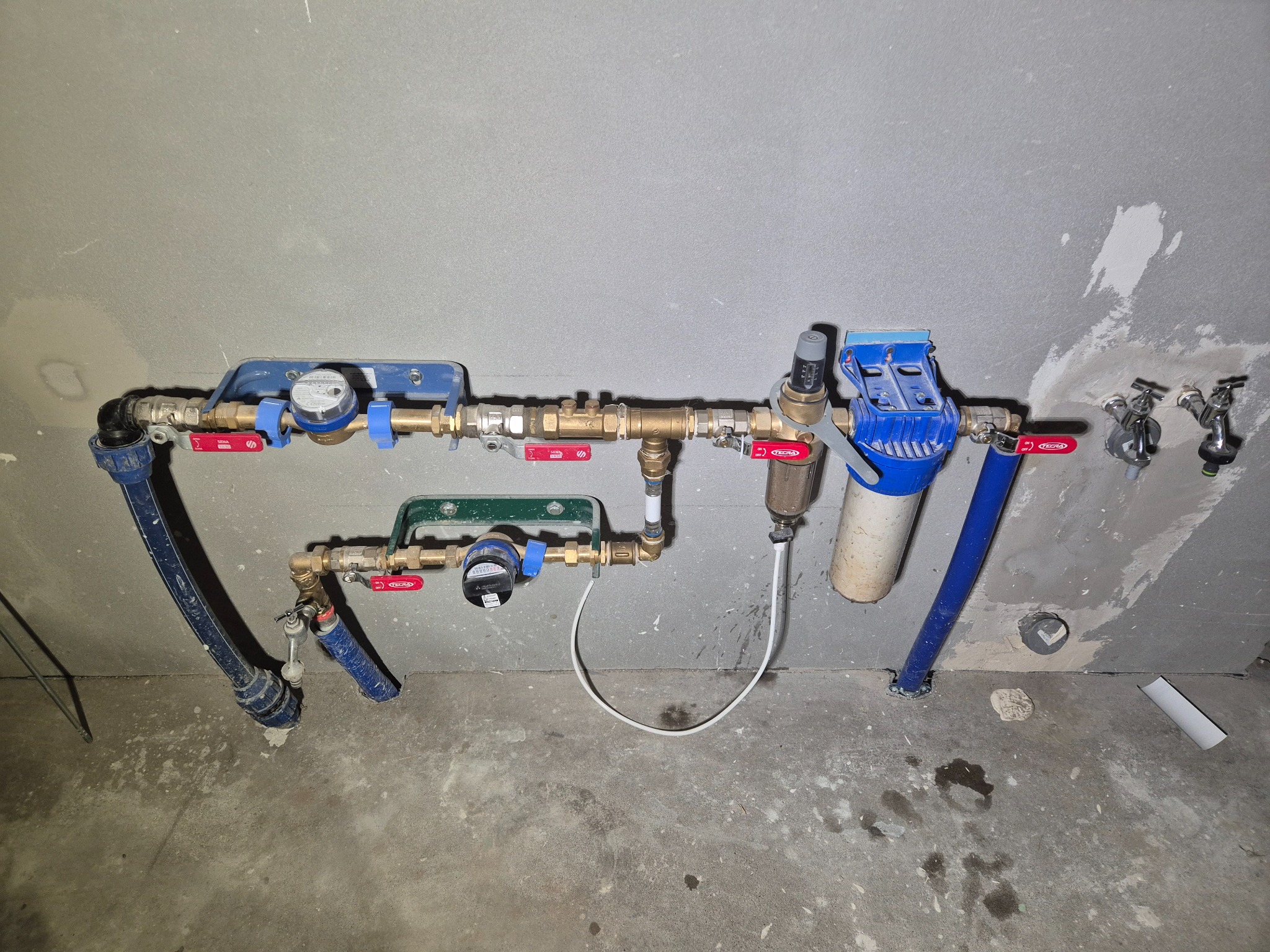 Dwa poziomy instalacji wodnej z filtrami i licznikami, zamontowane na surowej ścianie. Widoczne rury, zawory i przyłącza. Surowy stan wykończenia wnętrza.