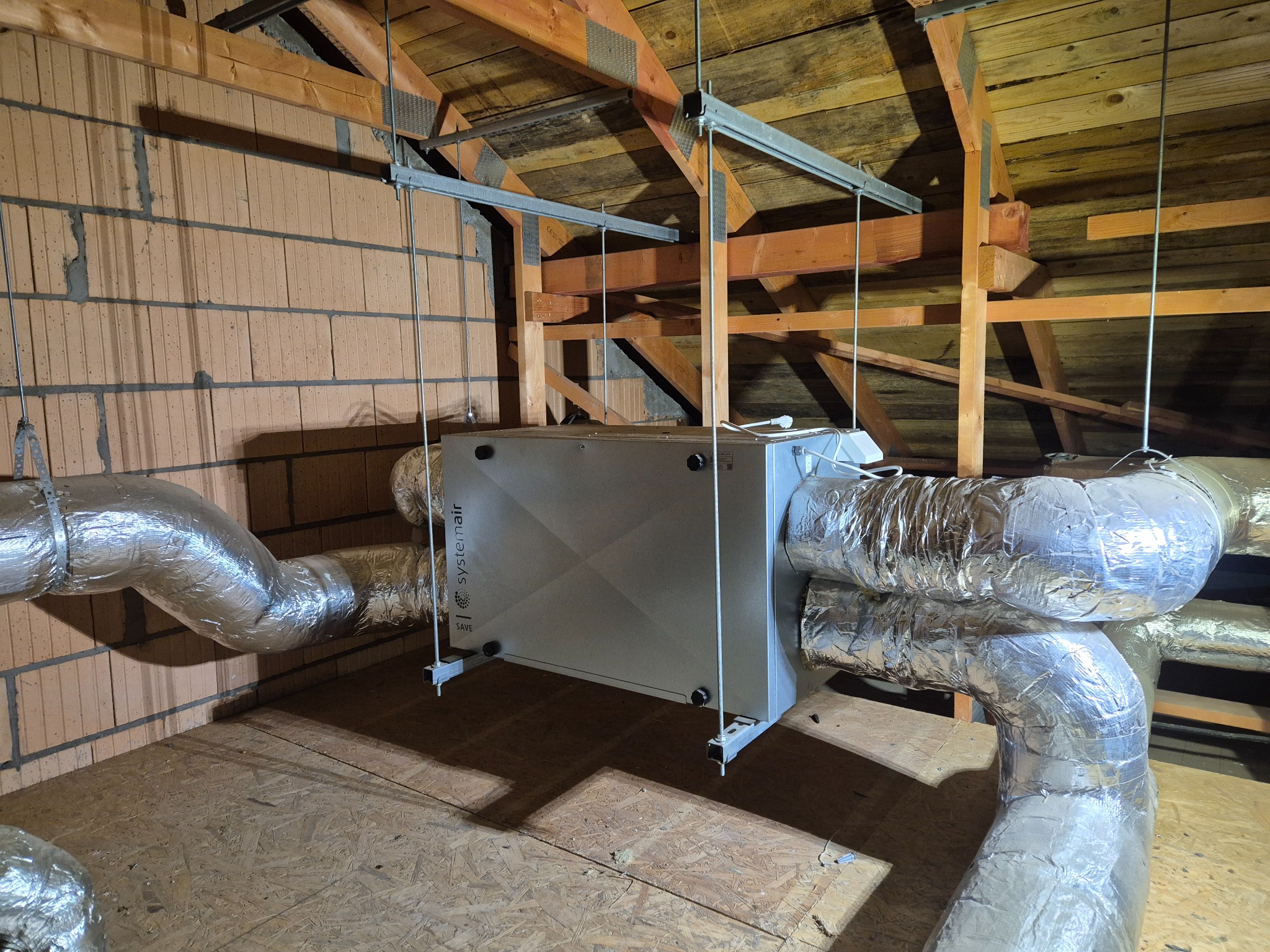 Centrala wentylacyjna Systemair SAVE zamontowana na poddaszu, z widocznymi przewodami wentylacyjnymi w izolacji aluminiowej, podwieszona do drewnianej konstrukcji dachu.