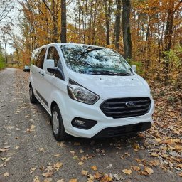 DBJ Transport - Biały Ford Transit na leśnej drodze, jesienne liście na ziemi. Widok z przodu, drzewa w tle. Auto gotowe do transportu.