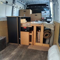 DBJ Transport - Wnętrze białego busa załadowane meblami, kartonami i zwiniętym dywanem, przygotowane do transportu. Widoczna szafka, stół i pudełka z elektroniką.
