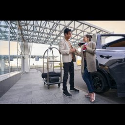 DBJ Transport - Elegancki mężczyzna w beżowej marynarce wita kobietę z paszportem przy czarnym samochodzie. W tle wózek bagażowy i przeszklony budynek lotniska.