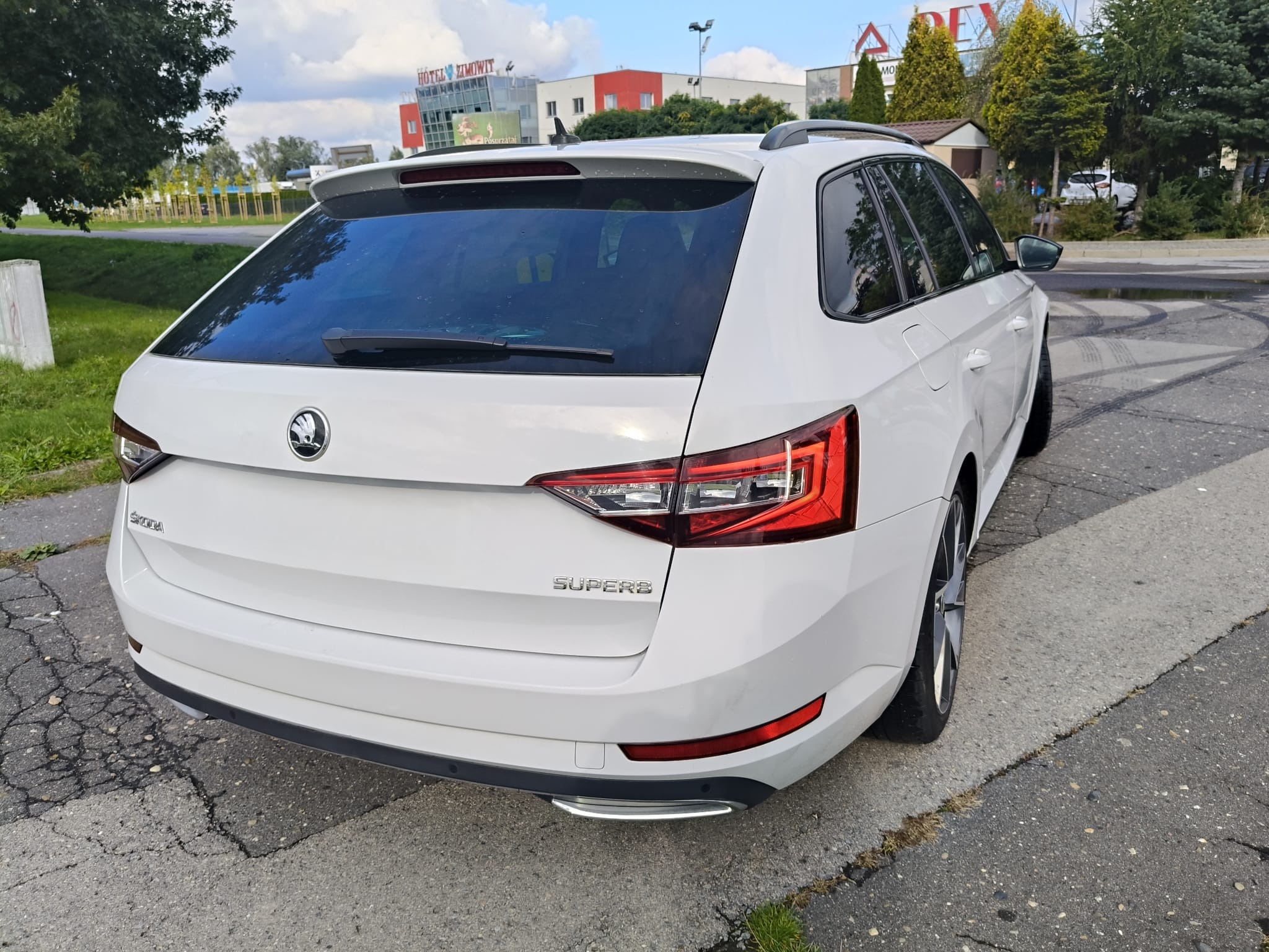 Biała Skoda Superb Combi z tyłu, zaparkowana na ulicy. Widoczne światła, logo i napis Superb. W tle budynki i drzewa. Auto czyste, zadbane, gotowe do drogi.
