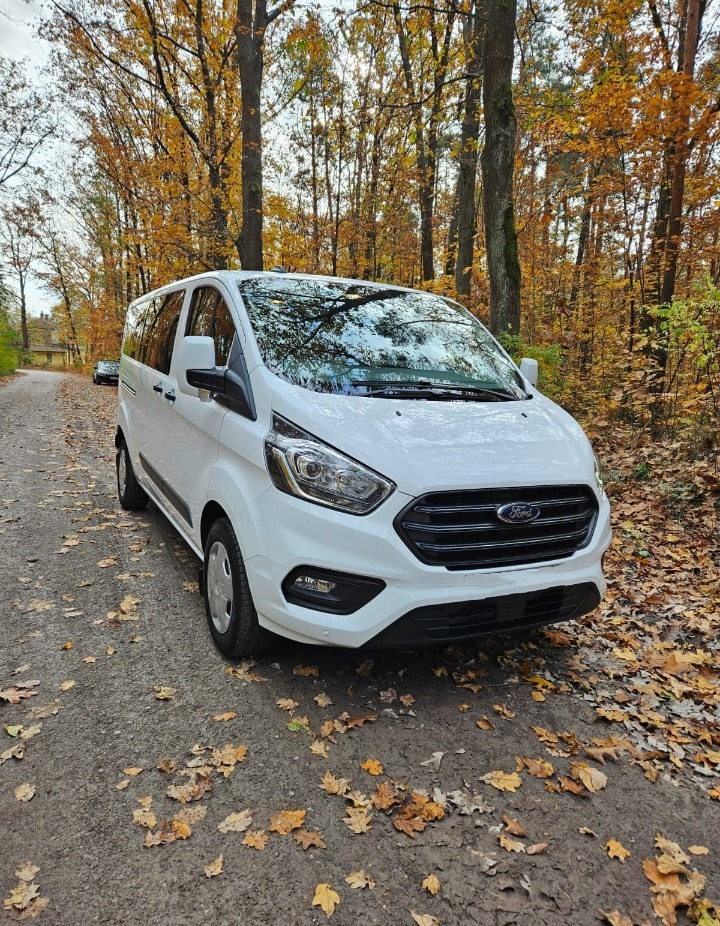Biały Ford Transit na leśnej drodze, jesienne liście na ziemi. Widok z przodu, drzewa w tle. Auto gotowe do transportu.