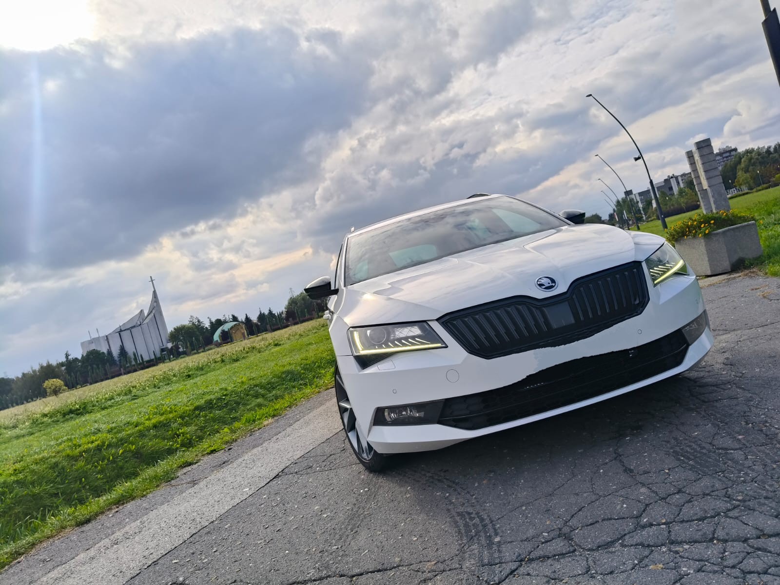 Biała Skoda Superb na tle kościoła i pochmurnego nieba, ujęcie z dołu, podkreślające nowoczesny design auta. Widoczne detale: czarny grill i reflektory.
