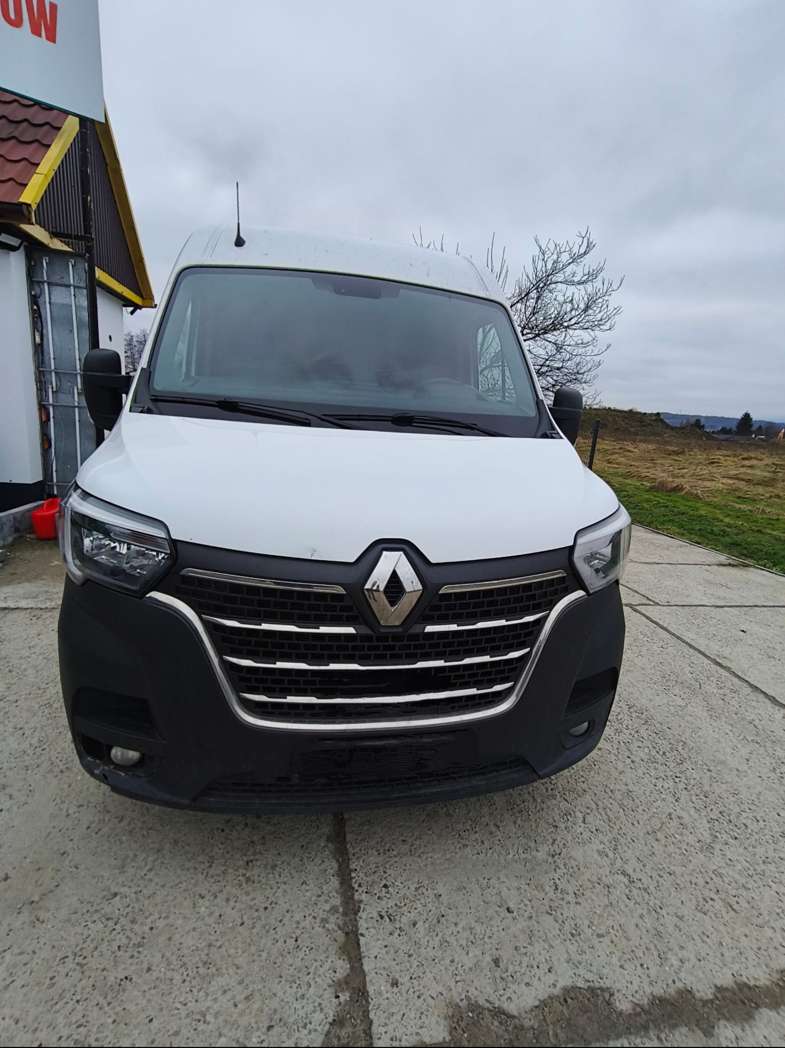 Biały Renault Master z widocznym grillem i reflektorami na tle pochmurnego nieba i budynku z żółtym dachem. Ujęcie z dołu podkreśla masywność pojazdu.