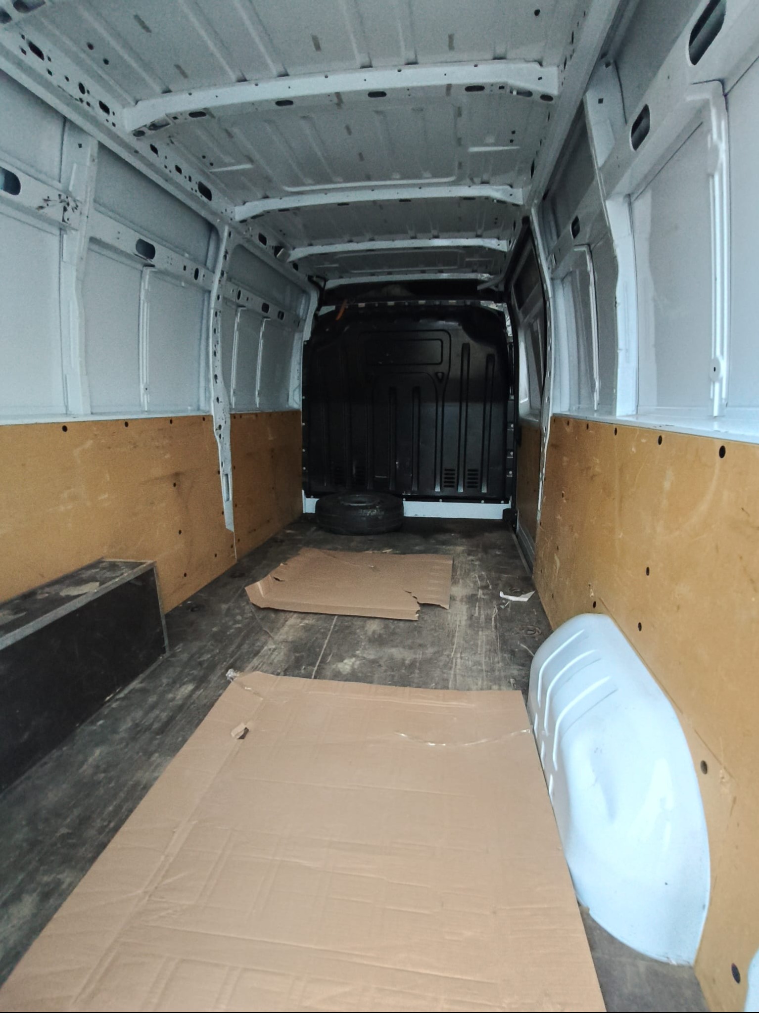 Wnętrze białego furgonetki z drewnianą okładziną ścian, przygotowane do transportu. Na podłodze leżą kartony, a w tle widoczna opona.