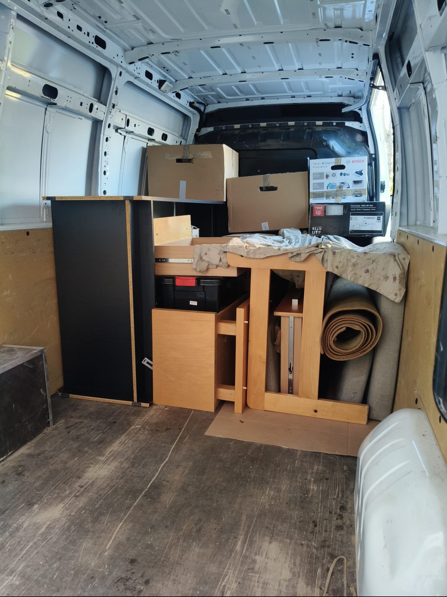 Wnętrze białego busa załadowane meblami, kartonami i zwiniętym dywanem, przygotowane do transportu. Widoczna szafka, stół i pudełka z elektroniką.