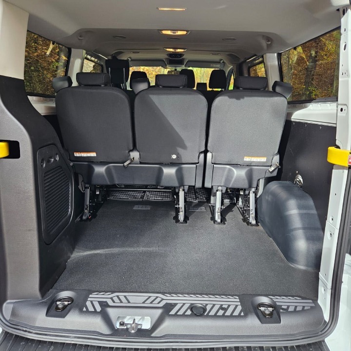 Wnętrze białego busa z trzema rzędami czarnych siedzeń, widok z tyłu pojazdu, czarna podłoga z szarymi wzorami, jasne oświetlenie wnętrza.